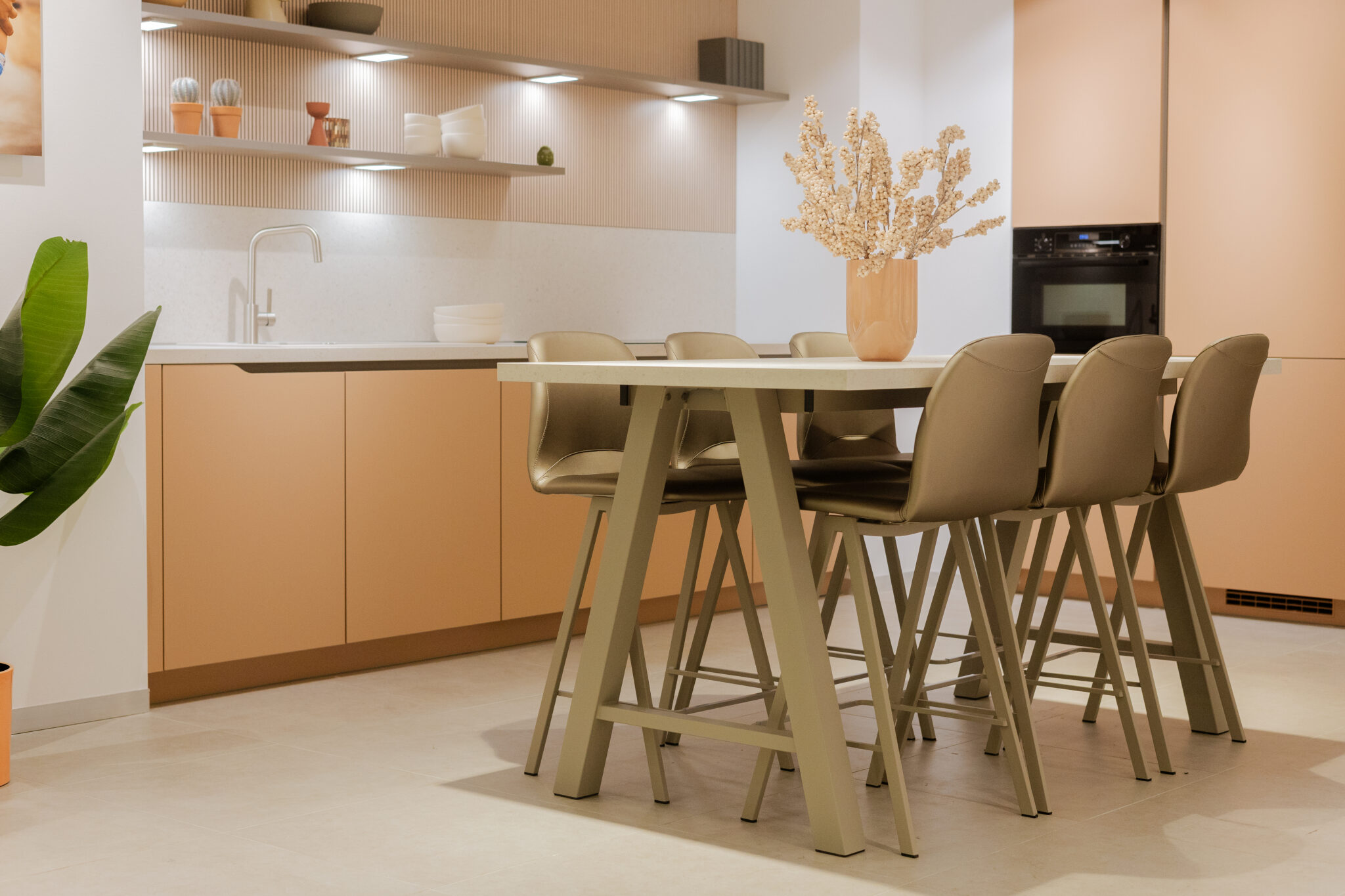 Cuisine moderne avec des placards couleur pêche, table à manger beige, chaises assorties et vase avec fleurs séchées sur la table.