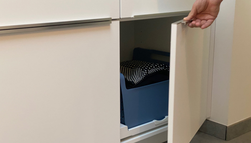 Armoire de cuisine blanche ouverte, révélant un bac de rangement bleu contenant des textiles noirs et blancs, avec une main tenant la porte.