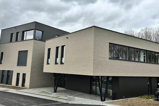 Bâtiment moderne à deux étages en briques beige avec grandes fenêtres et structure noire, sous un ciel nuageux.