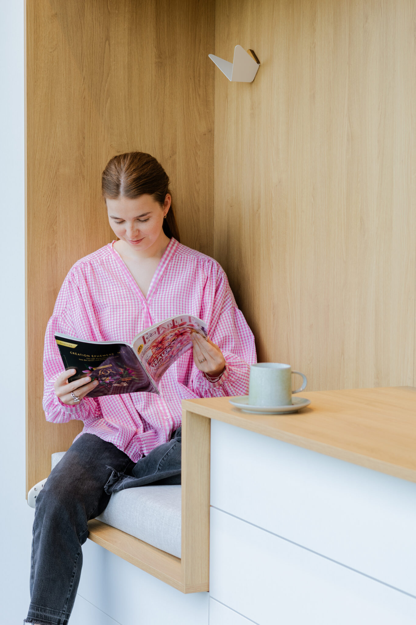 hall d’entrée Une personne en chemise rose lit un magazine assise sur un banc en bois clair, avec une tasse verte posée à côté.