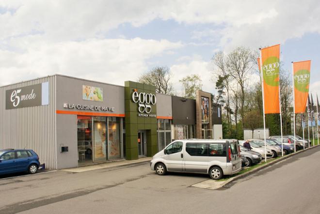 Façade d'un showroom èggo avec voitures garées devant, bâtiments gris et vert, et drapeaux orange alignés sur la droite.