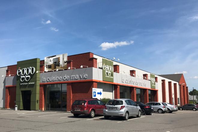 Façade d'un magasin èggo en briques rouges avec des voitures garées devant, sous un ciel bleu partiellement nuageux.