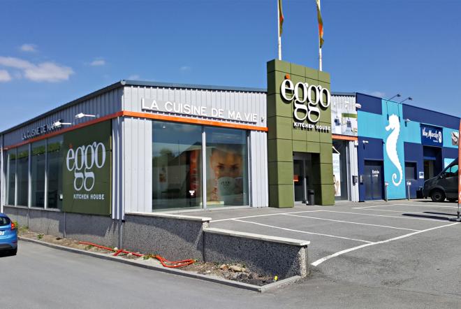 Magasin èggo avec façade grise et verte, logo visible, grandes vitrines et parking à l'avant sous un ciel bleu.