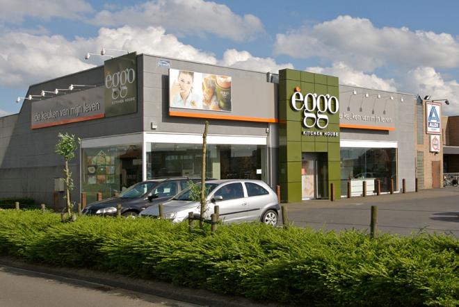 Façade d'un magasin èggo avec des voitures garées devant, enseigne verte et affiches publicitaires sur le mur gris.