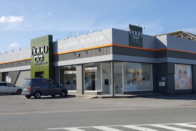 Façade d'un magasin èggo avec des panneaux gris et verts, une vitrine et une voiture garée devant sur le parking.