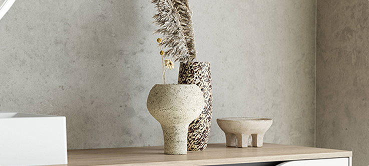 Deux vases en céramique beige et un bol sur pieds sont posés sur une surface en bois clair, devant un mur gris.