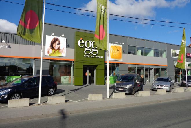Façade d'un magasin èggo avec des voitures garées devant, des drapeaux verts et un ciel bleu en arrière-plan.