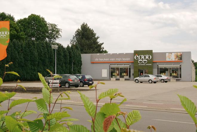 Façade d'un magasin èggo avec des voitures garées devant, entouré de haies vertes et d'un ciel nuageux.