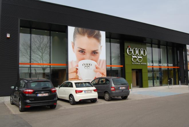 Façade d'un magasin èggo avec trois voitures garées devant, une grande affiche montrant une personne tenant une tasse de café.