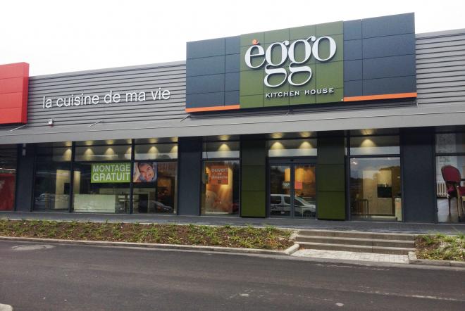 Façade d'un magasin èggo avec enseigne verte et noire, grandes vitrines et affiches publicitaires, sur un parking asphalté.