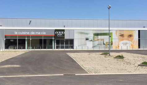 Façade d'un magasin èggo avec une grande affiche publicitaire, entourée d'un parking partiellement pavé et d'un ciel bleu.