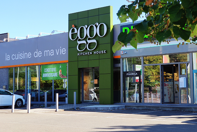 Façade d'un magasin èggo avec enseigne verte, vitrine, et panneaux publicitaires sous un ciel bleu.