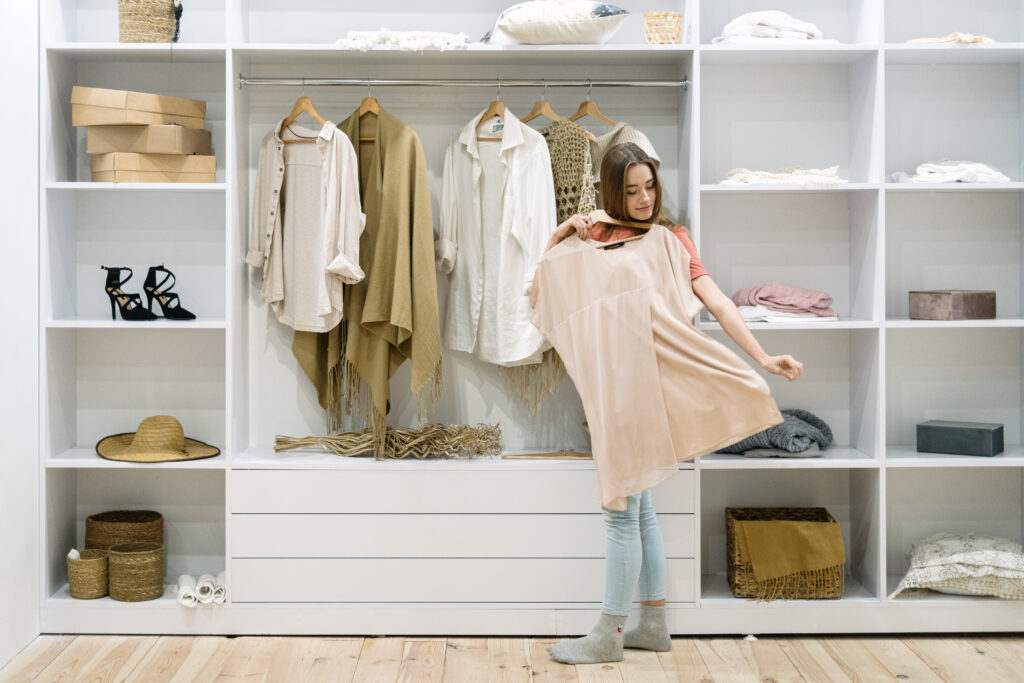Une femme tient une robe beige devant une garde-robe blanche avec vêtements suspendus et étagères remplies de boîtes, chaussures et paniers.