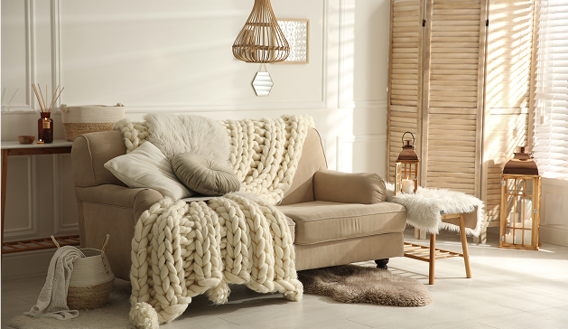 Un canapé beige recouvert d'un plaid en tricot crème est entouré de coussins, avec des lanternes et un paravent en bois à proximité.
