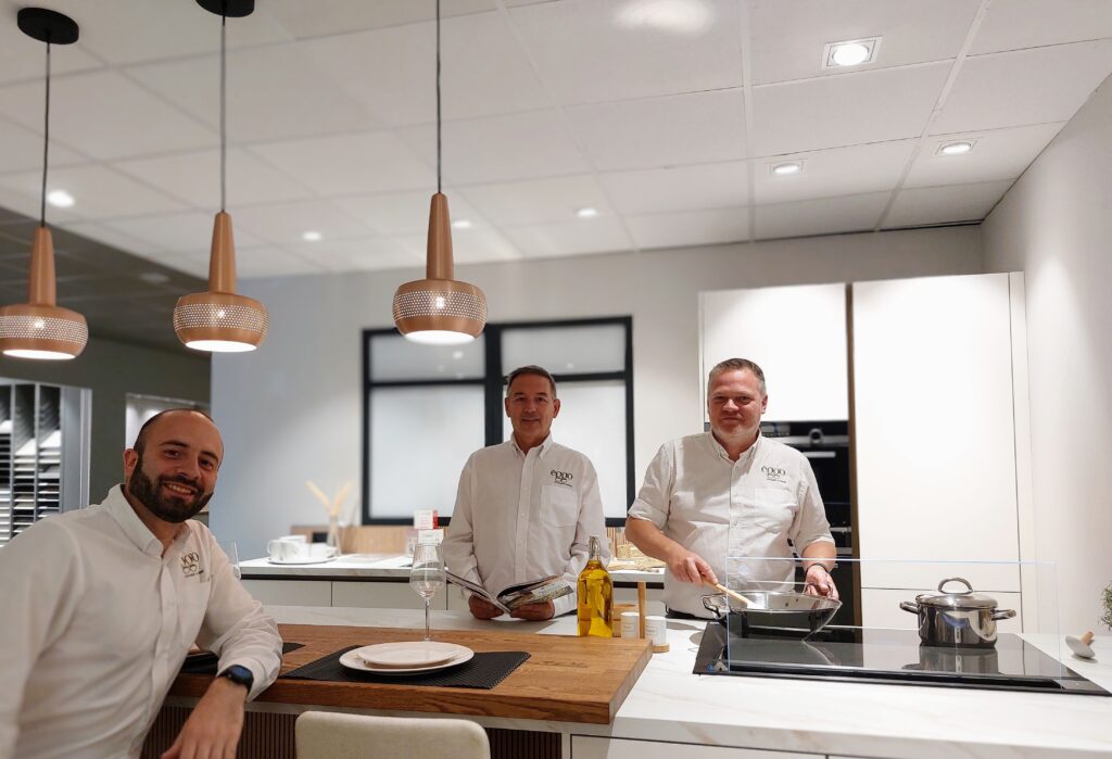 Trois hommes en chemise blanche dans une cuisine moderne avec des suspensions cuivrées, une plaque de cuisson et des ustensiles en acier inoxydable.