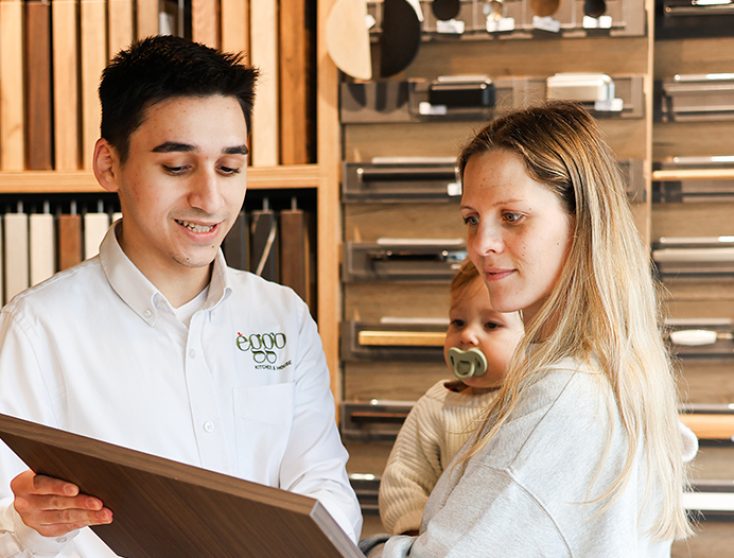 rencontre Un homme en chemise blanche montre un échantillon de bois à une femme tenant un enfant dans un showroom de cuisine.
